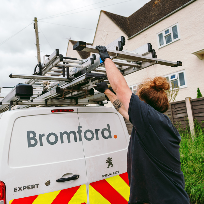 A Bromford engineer wearing gloves unloads a ladder from the roof rack of a white Bromford-branded Peugeot van parked on a residential street. The van has high-visibility chevrons on the rear, and houses are visible in the background.
