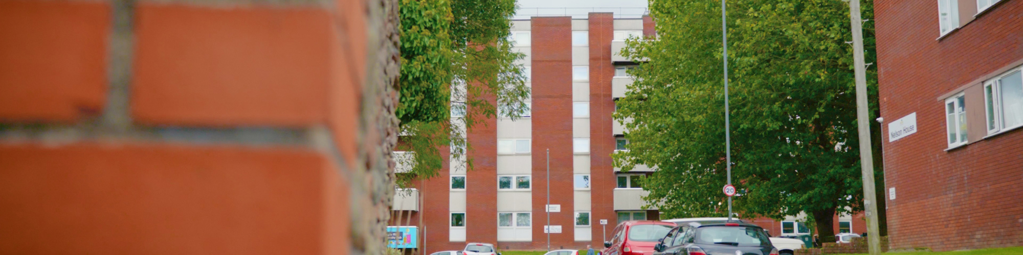 A street view framed on the left by a  close-up red brick wall corner. In the background, residential apartment buildings, green trees, and parked cars line a paved road under a cloudy sky.