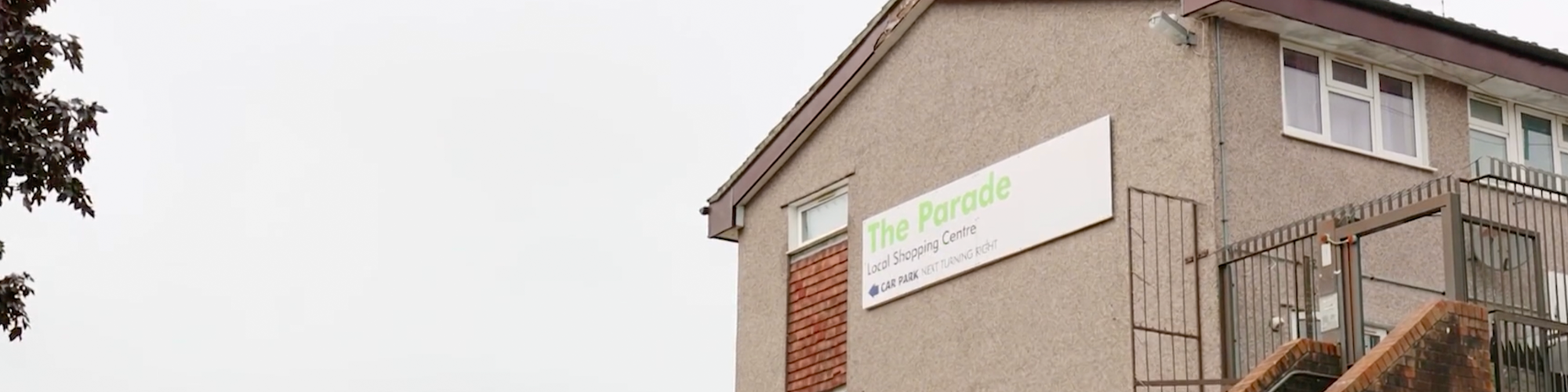 Exterior view of a multi-story building featuring a sign for "The Parade Local Shopping Centre" mounted on a grey wall, next to a red brick staircase, with a red post box and a Union Jack flag visible in the street below.