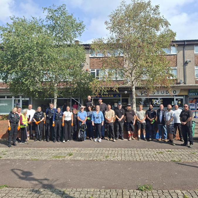 A large group of community volunteers, police officers, and tradespeople posing together outdoors in front of a row of shops and residential flats, standing along a paved walkway after a community cleanup event.