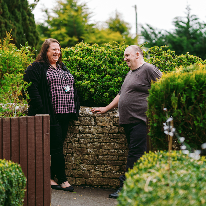 A Bromford neighbourhood coach and a  customer smiling and chatting next to a low stone wall in a residential garden setting.They are surrounded by hedges, trees, and garden foliage under bright daylight.