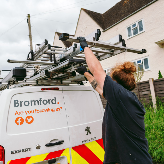 A tradesperson retrieving equipment from the roof rack of a white Bromford van that is parked on a residential street.