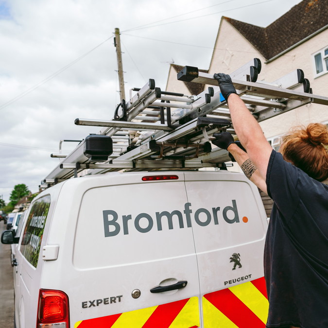 A tradesperson retrieving equipment from the roof rack of a white Bromford van that is parked on a residential street.