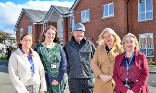 Five people stand side by side outdoors on a residential street, with modern red‑brick houses behind them. They are wearing smart and practical clothing, including coats and jackets, and are positioned facing the camera in daylight.
