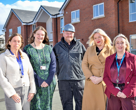 Five people stand side by side outdoors on a residential street, with modern red‑brick houses behind them. They are wearing smart and practical clothing, including coats and jackets, and are positioned facing the camera in daylight.