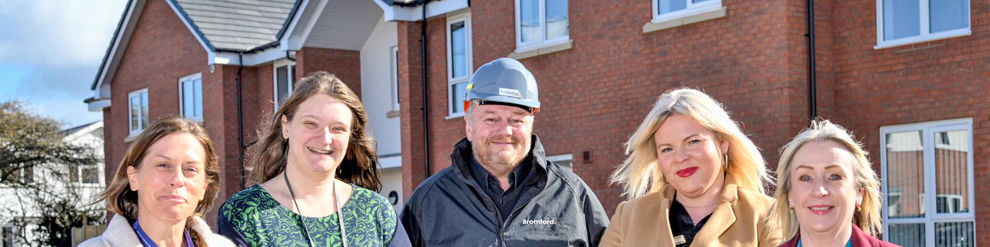 Five people stand side by side outdoors on a residential street, with modern red‑brick houses behind them. They are wearing smart and practical clothing, including coats and jackets, and are positioned facing the camera in daylight.