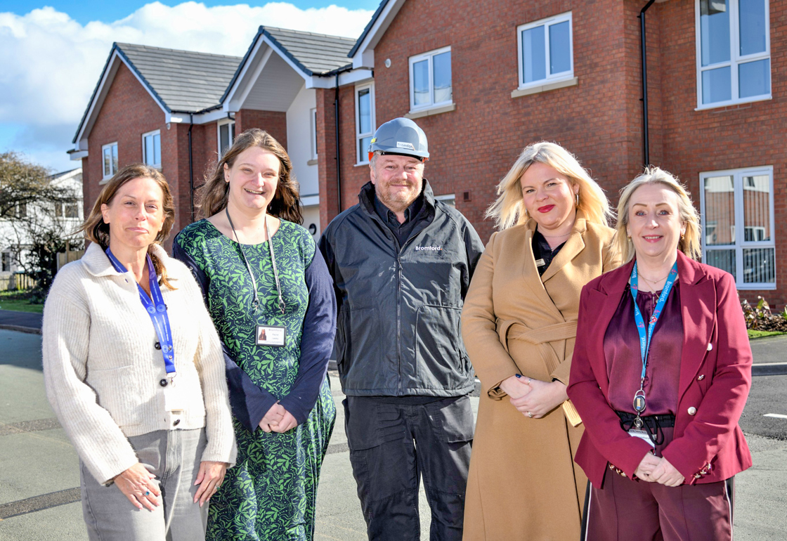 Five people stand side by side outdoors on a residential street, with modern red‑brick houses behind them. They are wearing smart and practical clothing, including coats and jackets, and are positioned facing the camera in daylight.