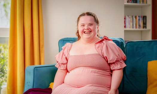 A woman sits on a sofa smiling while wearing a pink dress