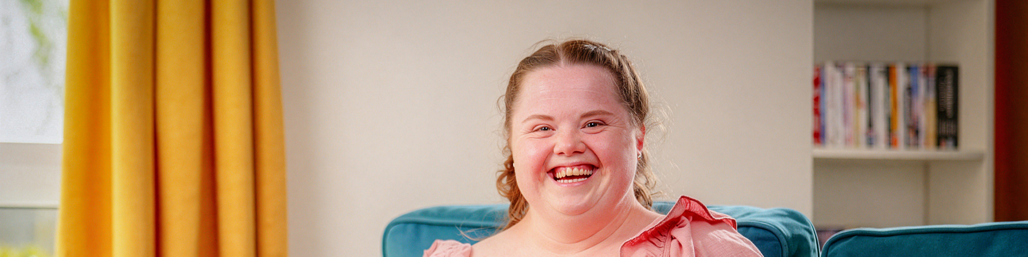 A woman sits on a sofa smiling while wearing a pink dress