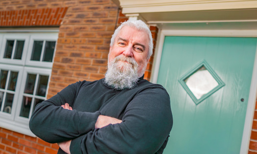 Person standing with arms crossed outside a brick house, wearing a dark long-sleeve top, positioned beside a green front door and a white-framed window.