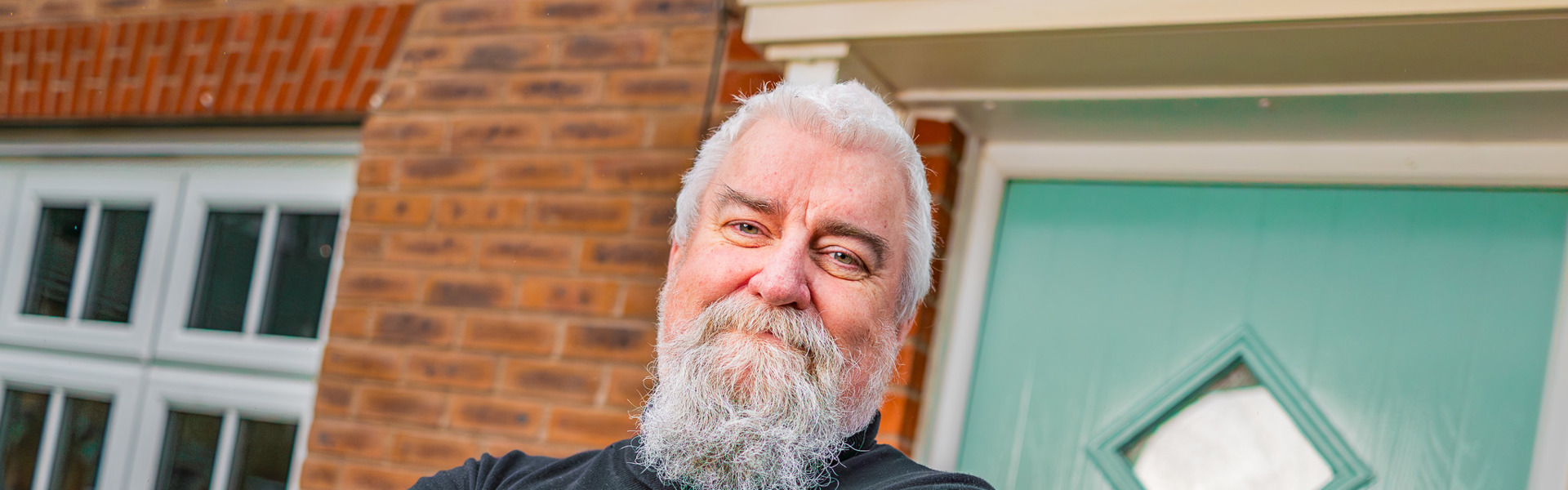 Person standing with arms crossed outside a brick house, wearing a dark long-sleeve top, positioned beside a green front door and a white-framed window.