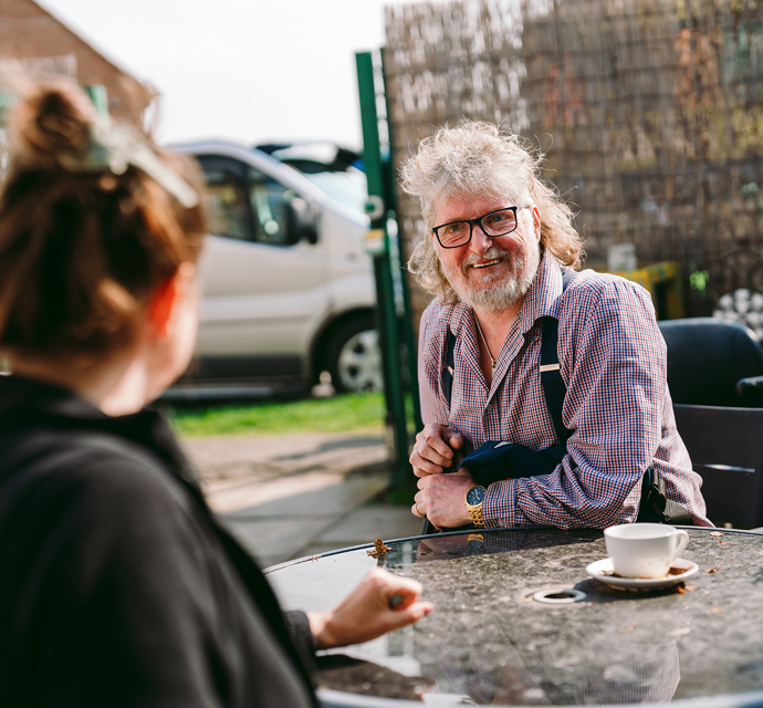 A bromford customer talking to their neighbourhood coach at a table outside. In the background, a residential driveway and a white van are visible.