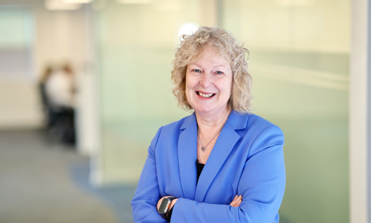 A smiling person with curly blonde hair wearing a blue blazer, standing with arms crossed in a bright modern office corridor.