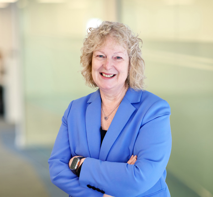 A smiling person with curly blonde hair wearing a blue blazer, standing with arms crossed in a bright modern office corridor.